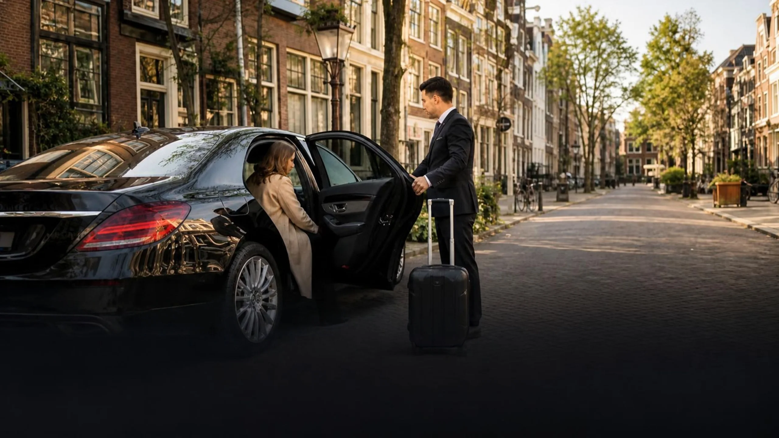 A driver holding the door of a black Mercedes for a traveller with a suitcase on a street lined with old town houses